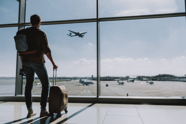 Man is standing with luggage near window in waiting room. He is glancing at plane taking off outside. Copy space in right side